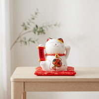 Maneki-neko porte-bonheur en céramique émaillée, deux pattes levées, posé sur un coussin brocart dans un intérieur clair
